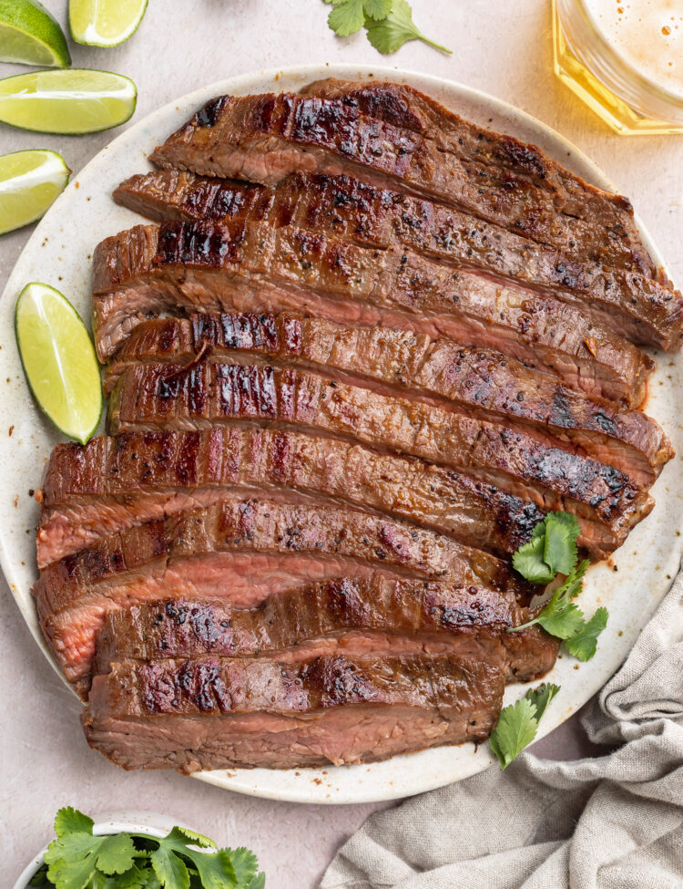 Top-down view of a sliced, sous-vide cooked flank steak on a white plate surrounded by lime wedges and other garnish.