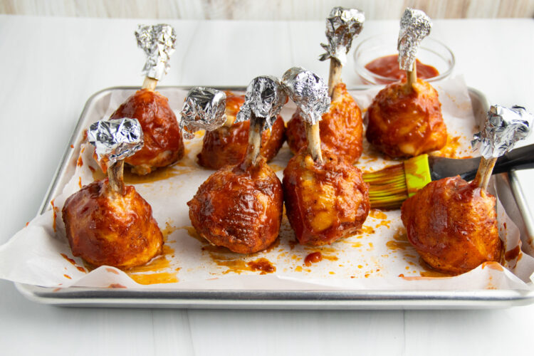 Lollipop chicken legs, with exposed bones covered by aluminum foil, standing upright on a baking sheet lined with parchment paper. The chicken is generously coated in BBQ sauce.