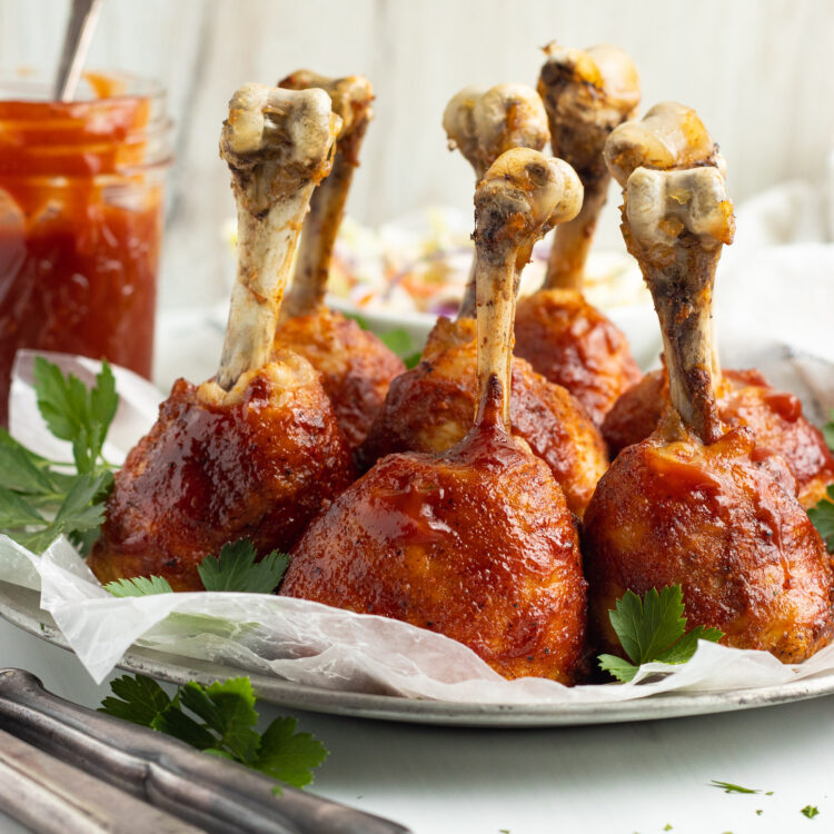 Side view of lollipop chicken legs coated in bbq sauce, standing upright on a baking sheet lined with parchment paper.