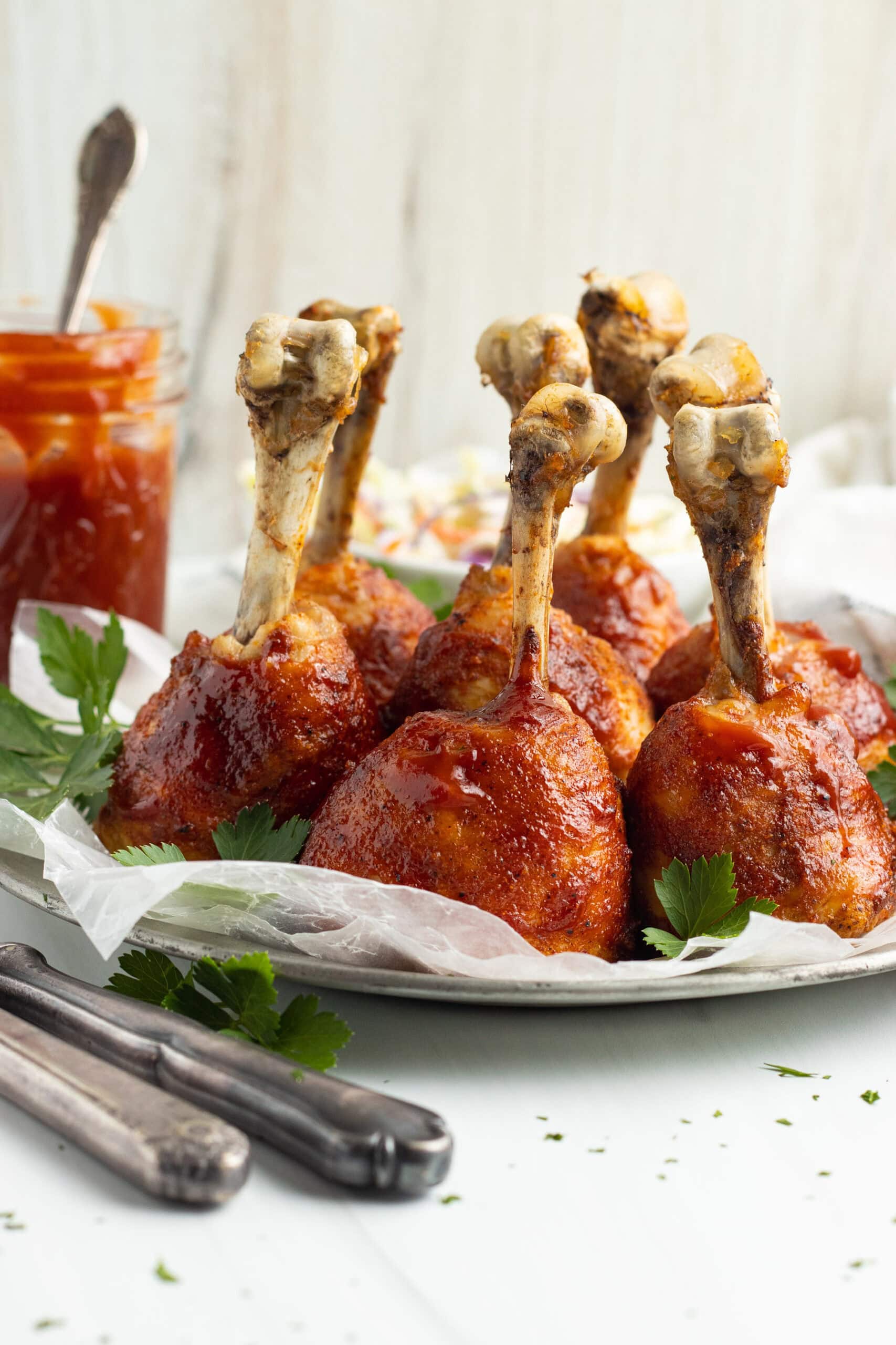 Side view of lollipop chicken legs coated in bbq sauce, standing upright on a baking sheet lined with parchment paper.