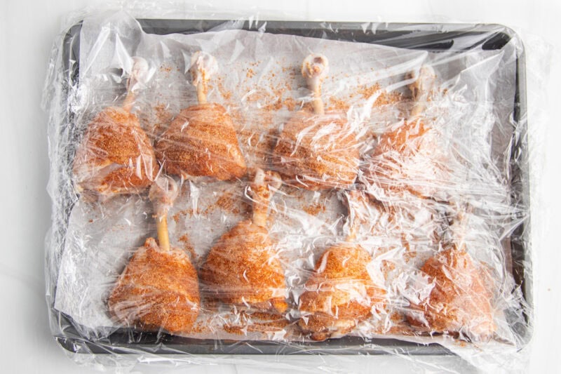 Dry rub seasoned drumsticks laying on a baking sheet lined with aluminum foil, covered with plastic wrap.
