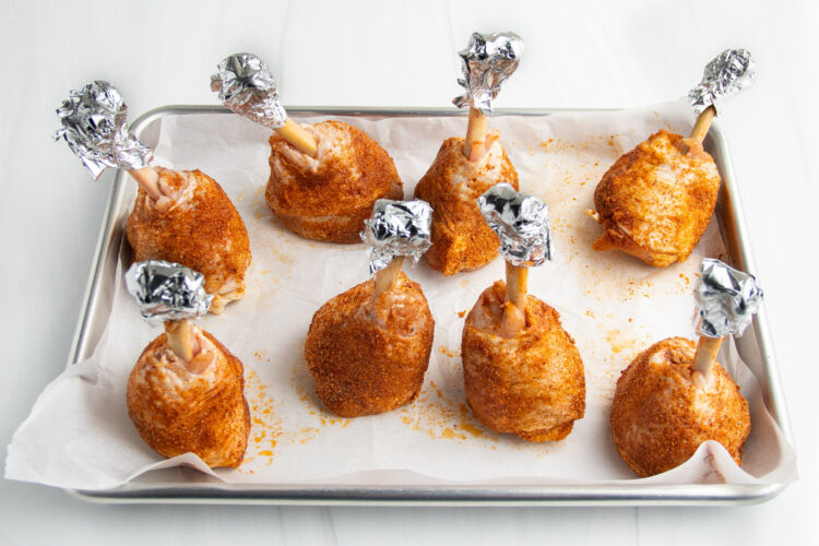 Lollipop chicken legs, with exposed bones covered by aluminum foil, standing upright on a baking sheet lined with parchment paper.