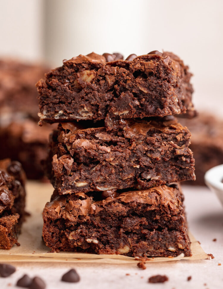 Three thick, rich, chocolatey lactation brownies stacked on top of each other, with chocolate chips and crumbs on the counter around them.