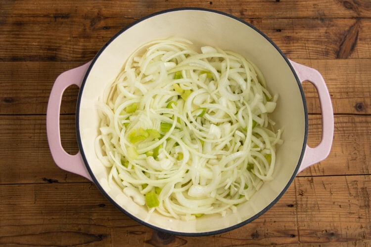 Sliced onions and chopped celery in a large pot on a wooden tabletop.