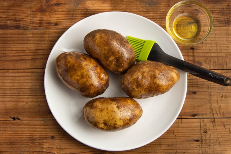 Olive oil being brushed onto 4 russet potatoes with a basting brush.
