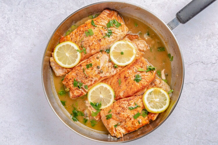 Salmon piccata in a large silver skillet with lemon coins and parsley to garnish.