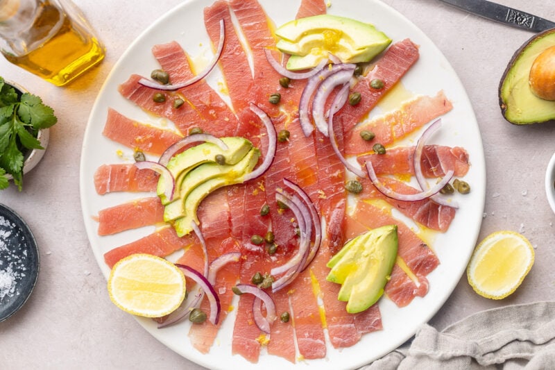 Tuna carpaccio arranged on a large white platter, topped with slices of avocado and red onion, with dots of capers.