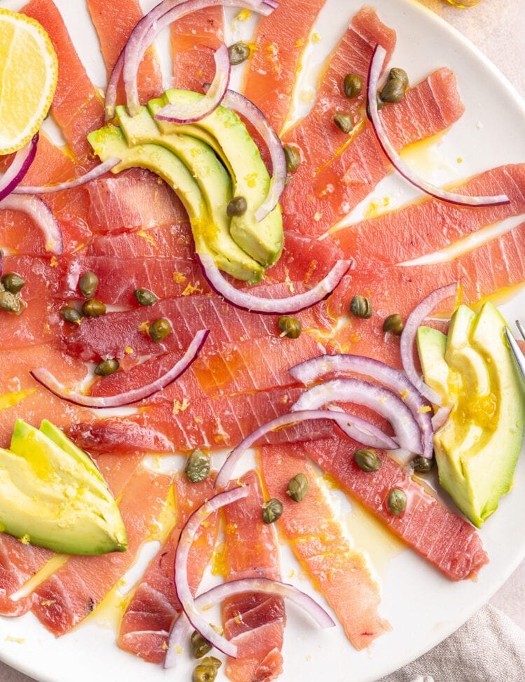 Tuna carpaccio arranged on a large white platter, topped with slices of avocado and red onion, with dots of capers.