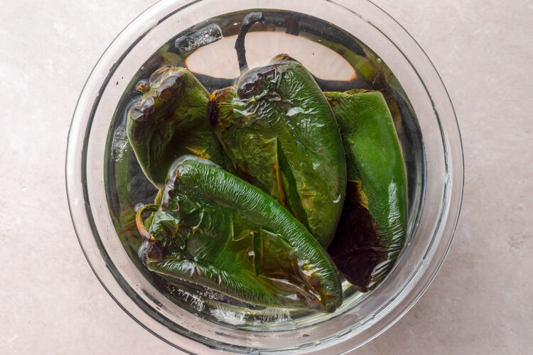 Charred and blistered poblano peppers in a bowl of cold water.