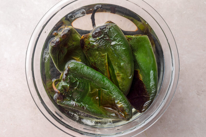 Charred and blistered poblano peppers in a bowl of cold water.