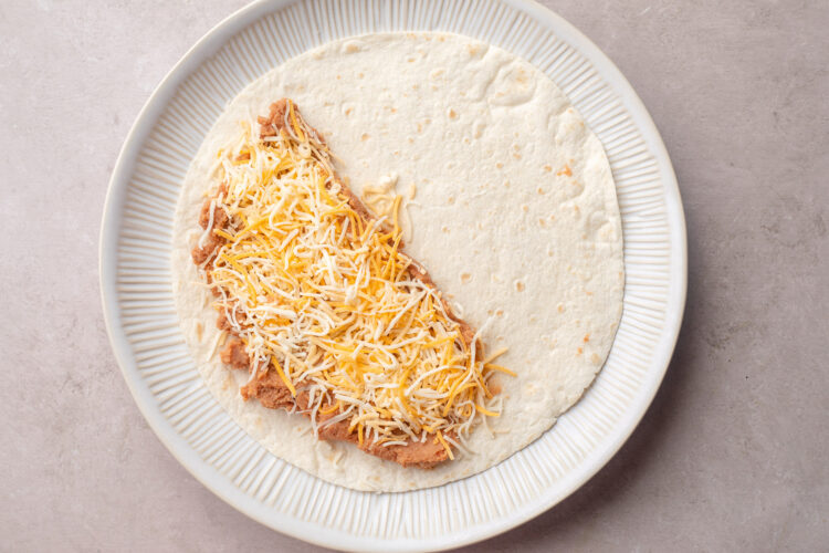 Refried beans and shredded cheese on one half of a 10-inch flour tortilla on a plate.