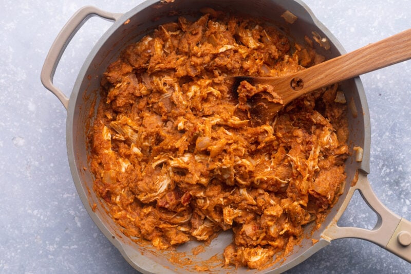 Shredded chicken, refried beans, salsa, taco seasoning, and onions in a large skillet with a wooden spoon.