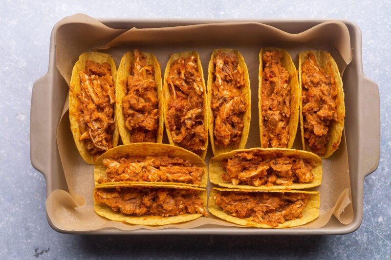 Crunchy taco shells upright in a baking pan and filled with shredded chicken mixture.