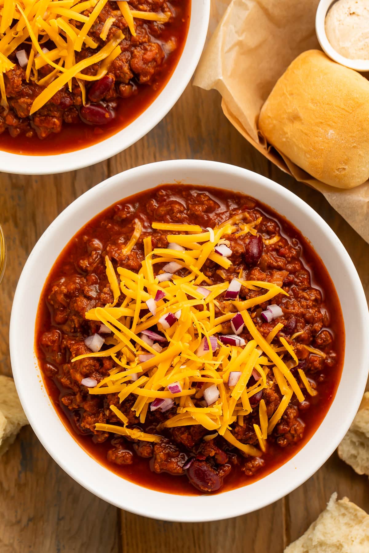 Overhead view of a bowl of dark red Texas Roadhouse chili topped with orange shreds of cheddar cheese and diced red onion on a dark tabletop.