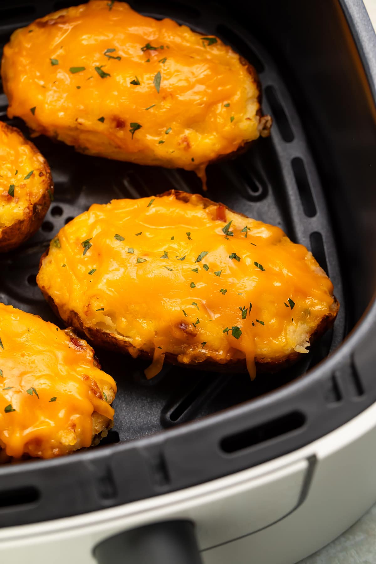 Twice baked potatoes in an air fryer basket. The potatoes are topped with a layer of melted cheddar and a garnish of chives.