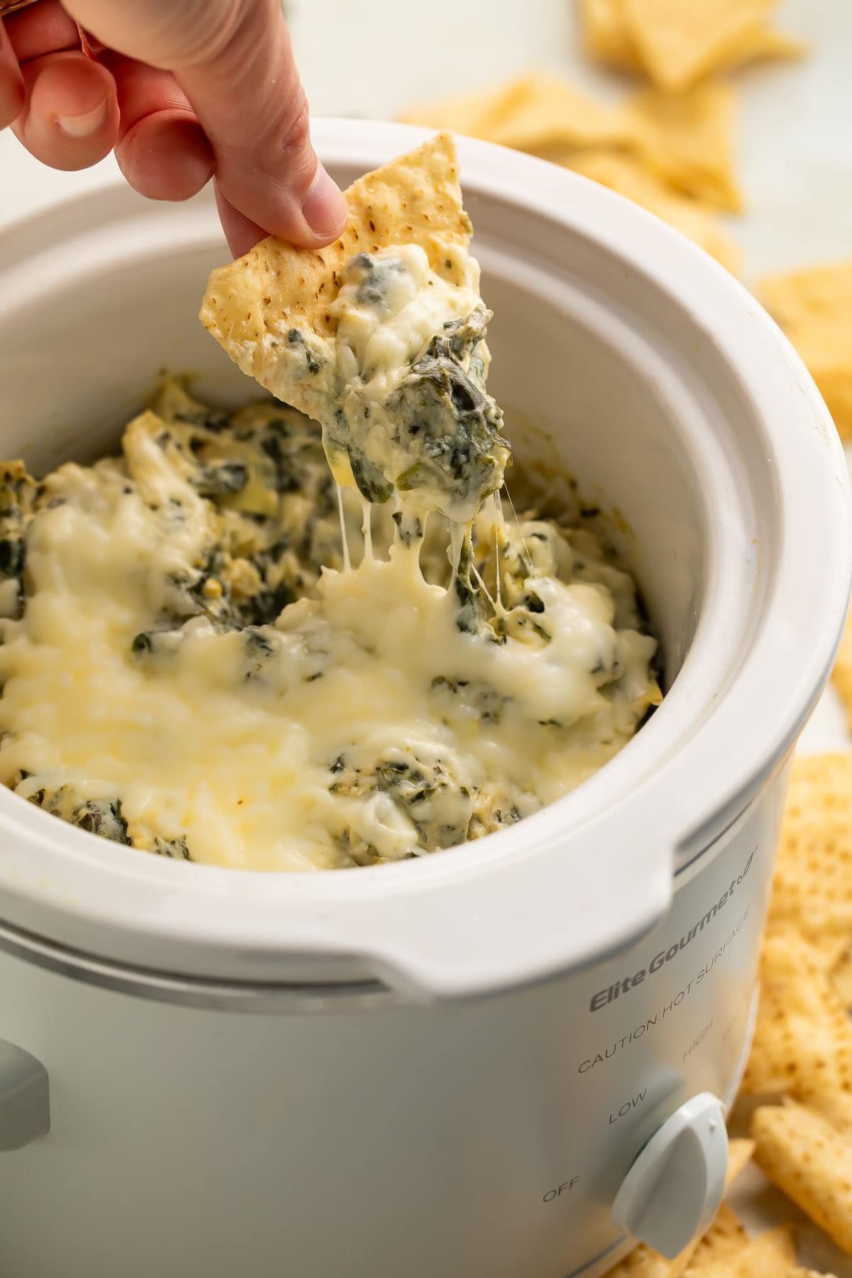 A woman holds a tortilla chip as she uses it to scoop creamy, cheesy spinach artichoke dip out of a white Crockpot slow cooker.