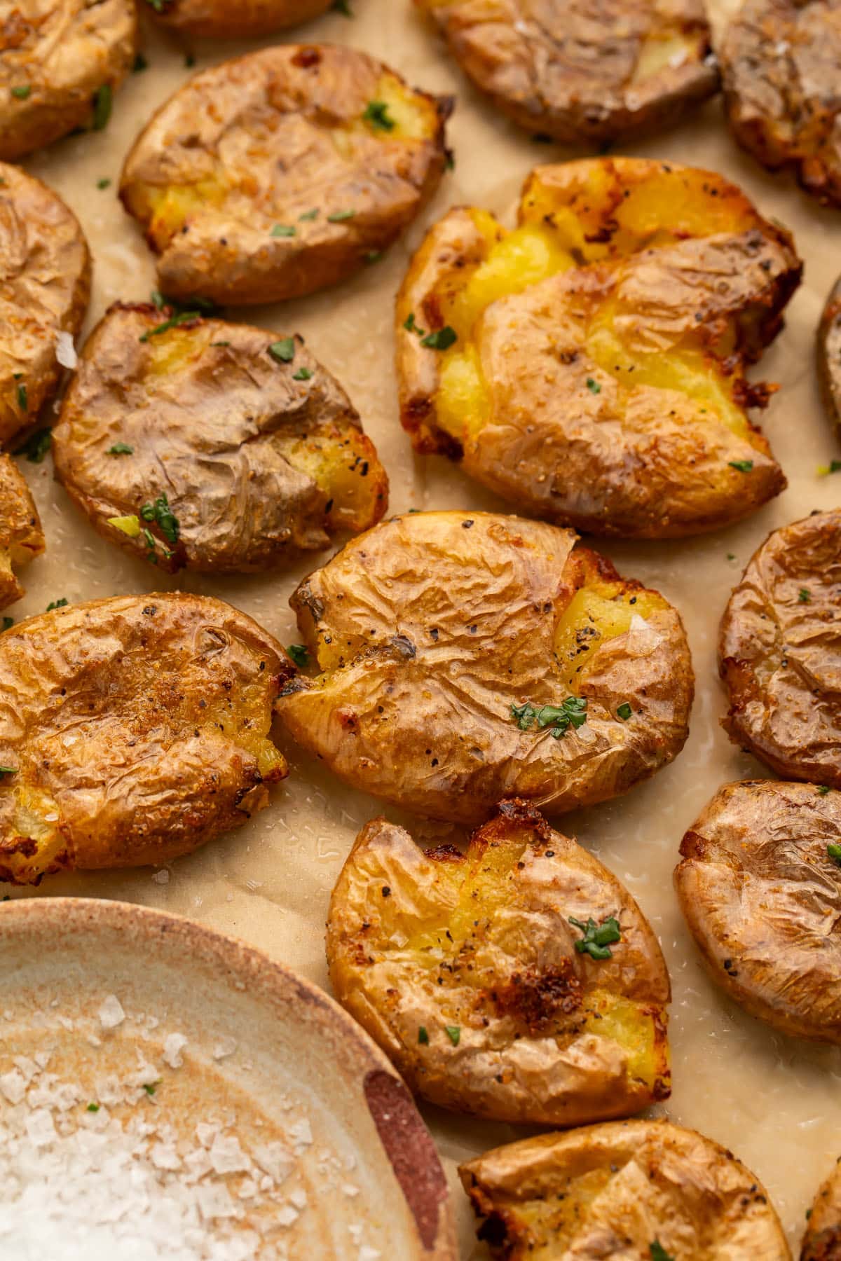 Multiple smashed potatoes, cooked in the air fryer, arranged on a sheet of parchment paper next to a small dish of salt.