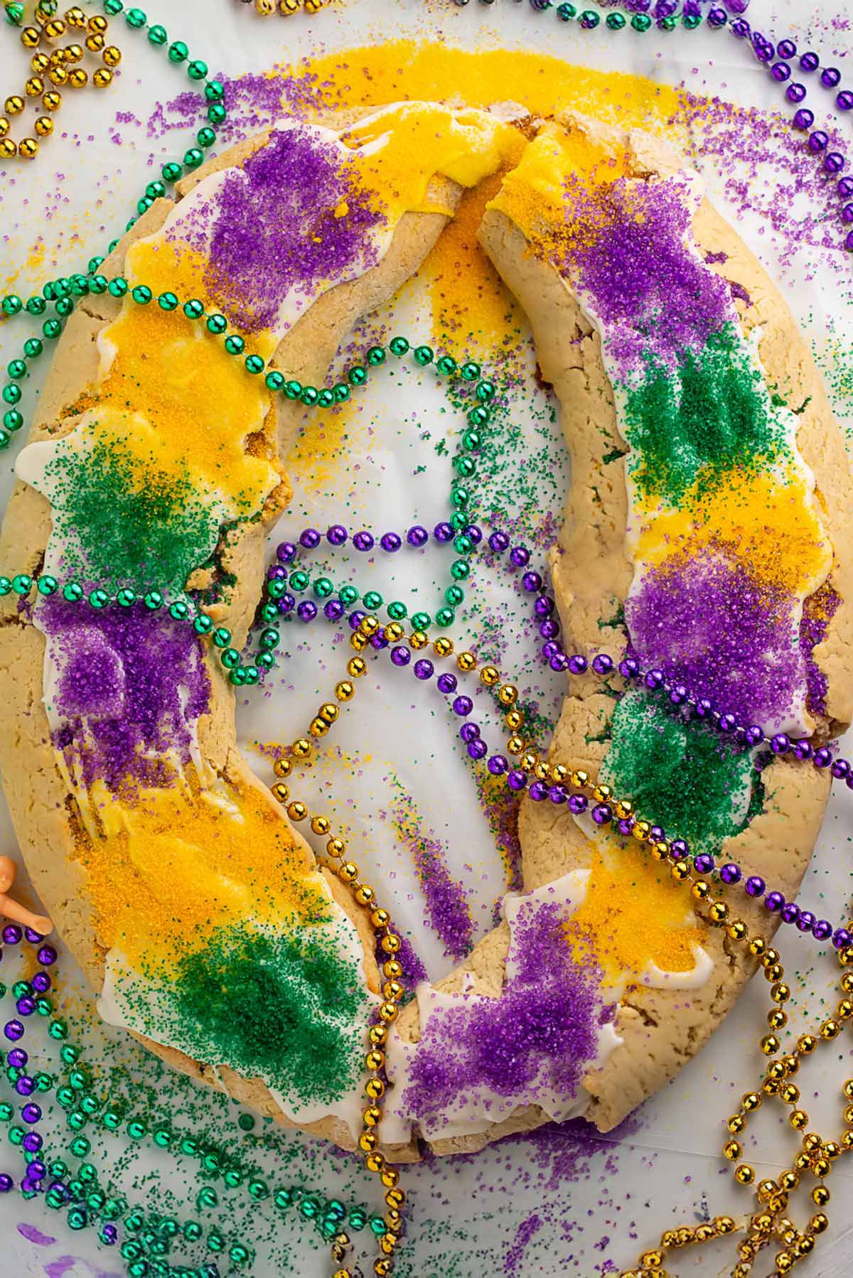 A large oval-shaped gluten free king cake topped with purple, yellow, and green sanding sugar and surrounding by purple, gold, and green Mardi Gras bead necklaces.