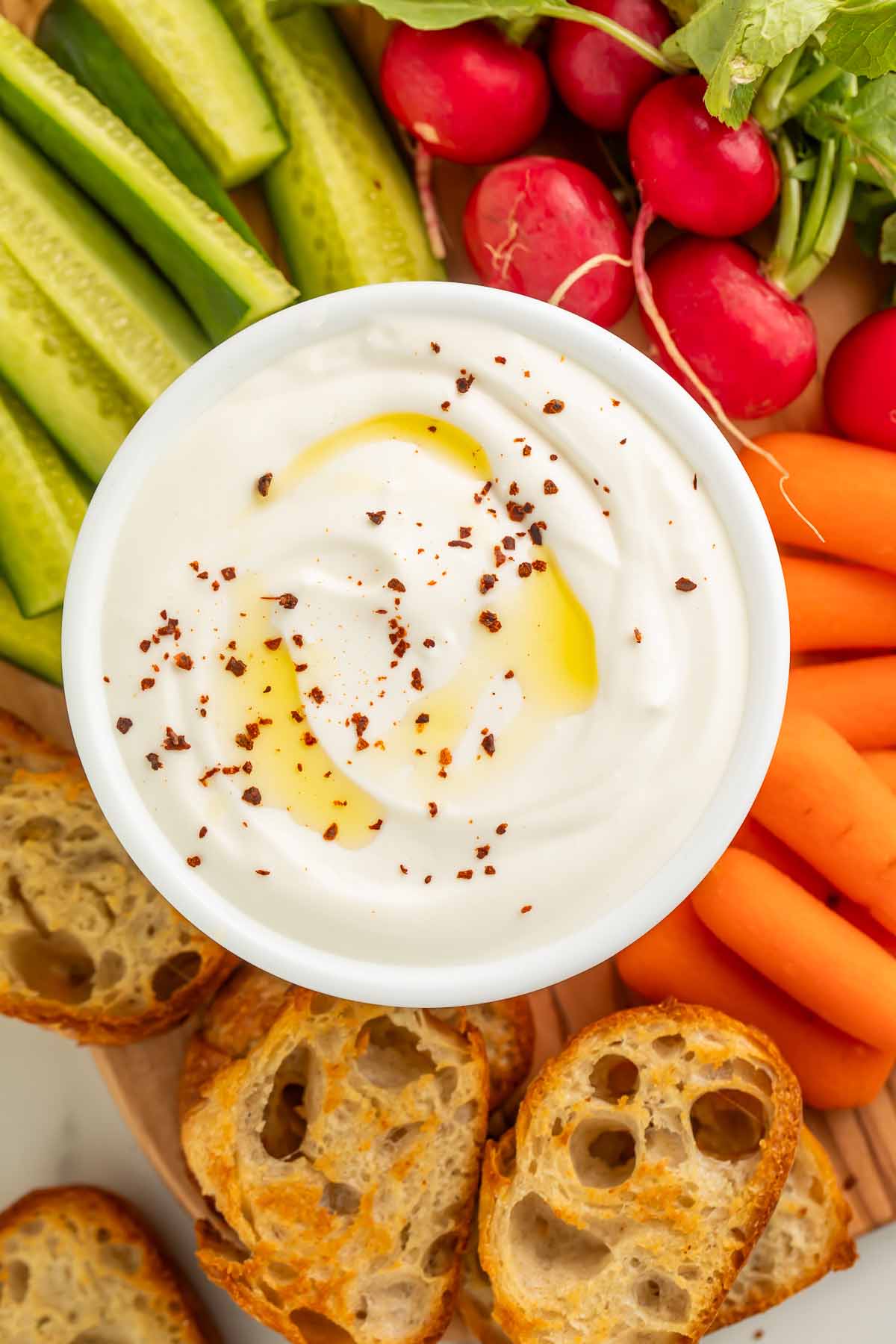 A bowl of savory whipped cottage cheese resting on a wooden board, surrounded by crostini, pickles, carrot sticks, and radishes.