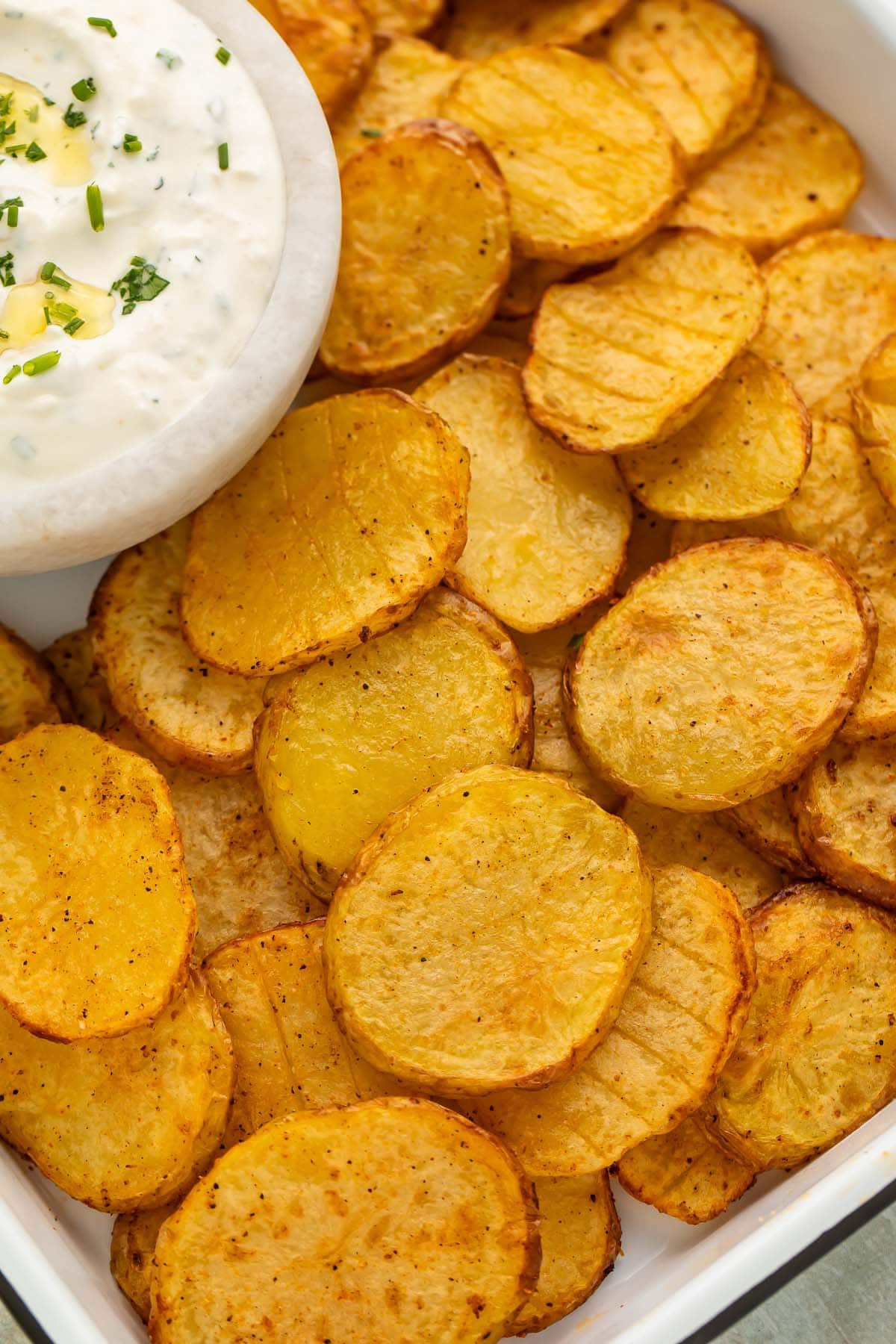 Close-up of thinly sliced potatoes, cooked in the air fryer, on a tray with a bowl of creamy dipping sauce.