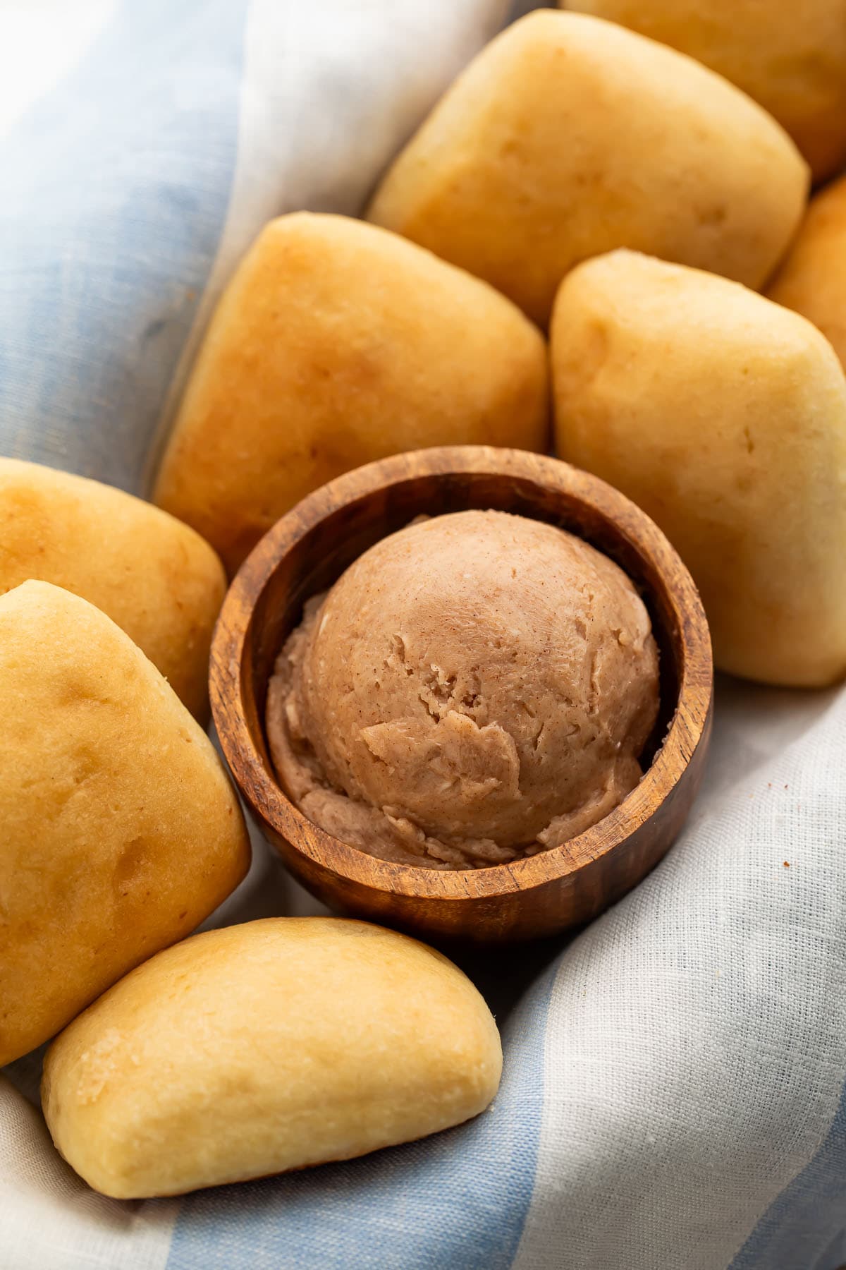 A small wooden bowl containing a scoop of rich brown Texas Roadhouse cinnamon honey butter nestled in a basket with golden yeast rolls.