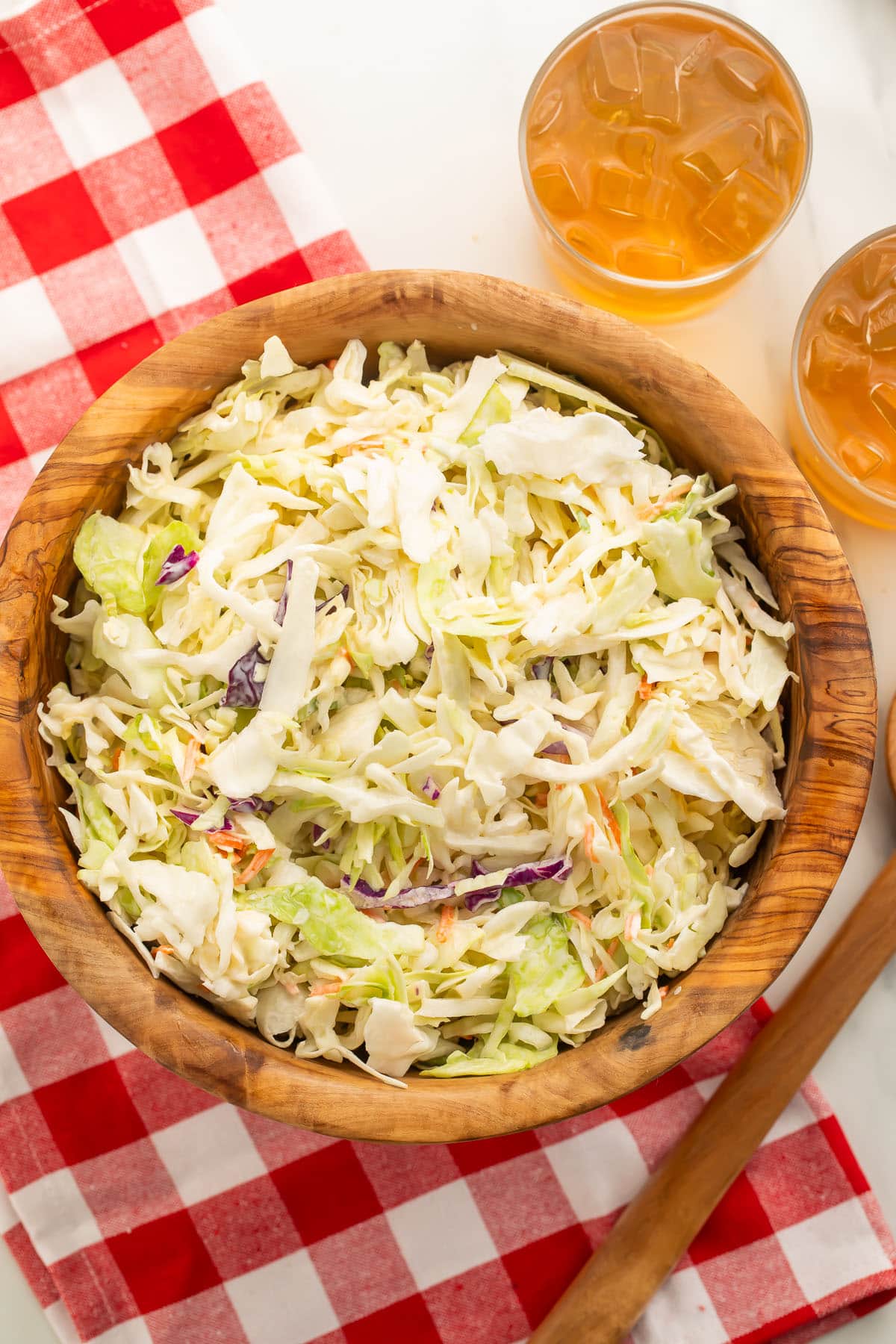 A wooden bowl containing vegan coleslaw sits on a table partially covered by a red and white checkerboard tablecloth.