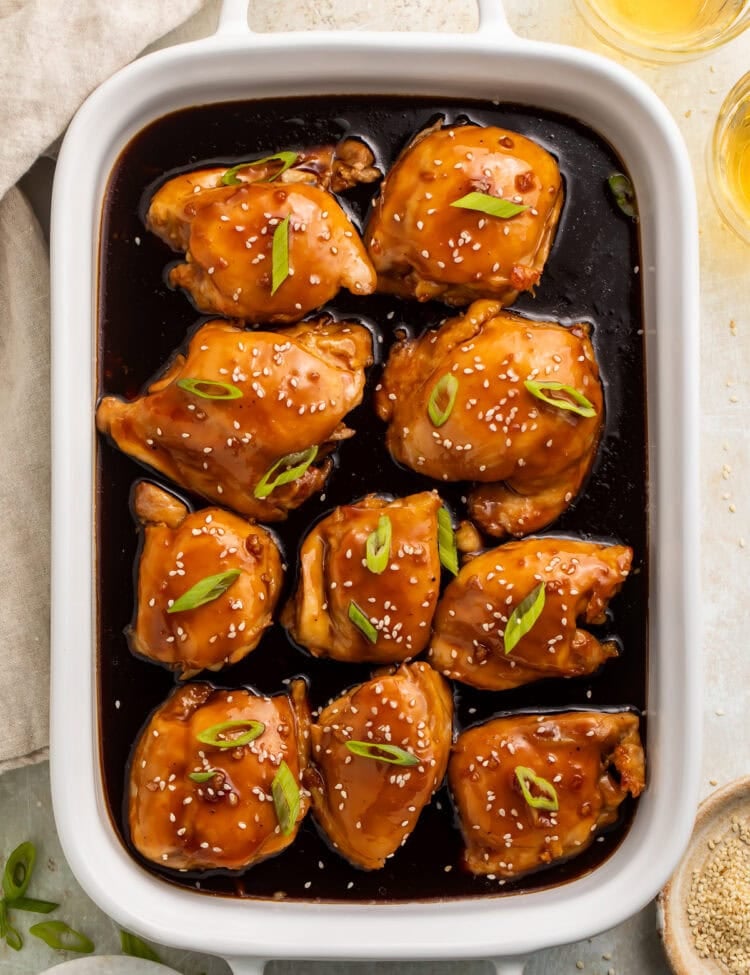 White baking dish with teriyaki chicken thighs and sauce, topped with sliced green onions and sesame seeds.