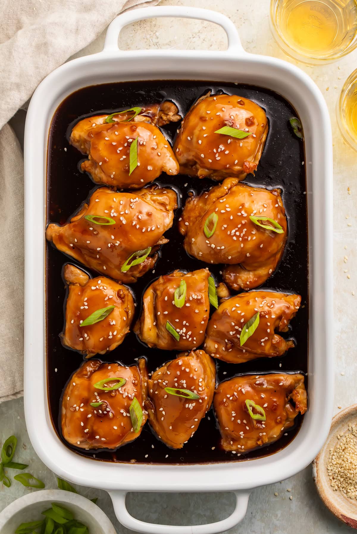 White baking dish with teriyaki chicken thighs and sauce, topped with sliced green onions and sesame seeds. 