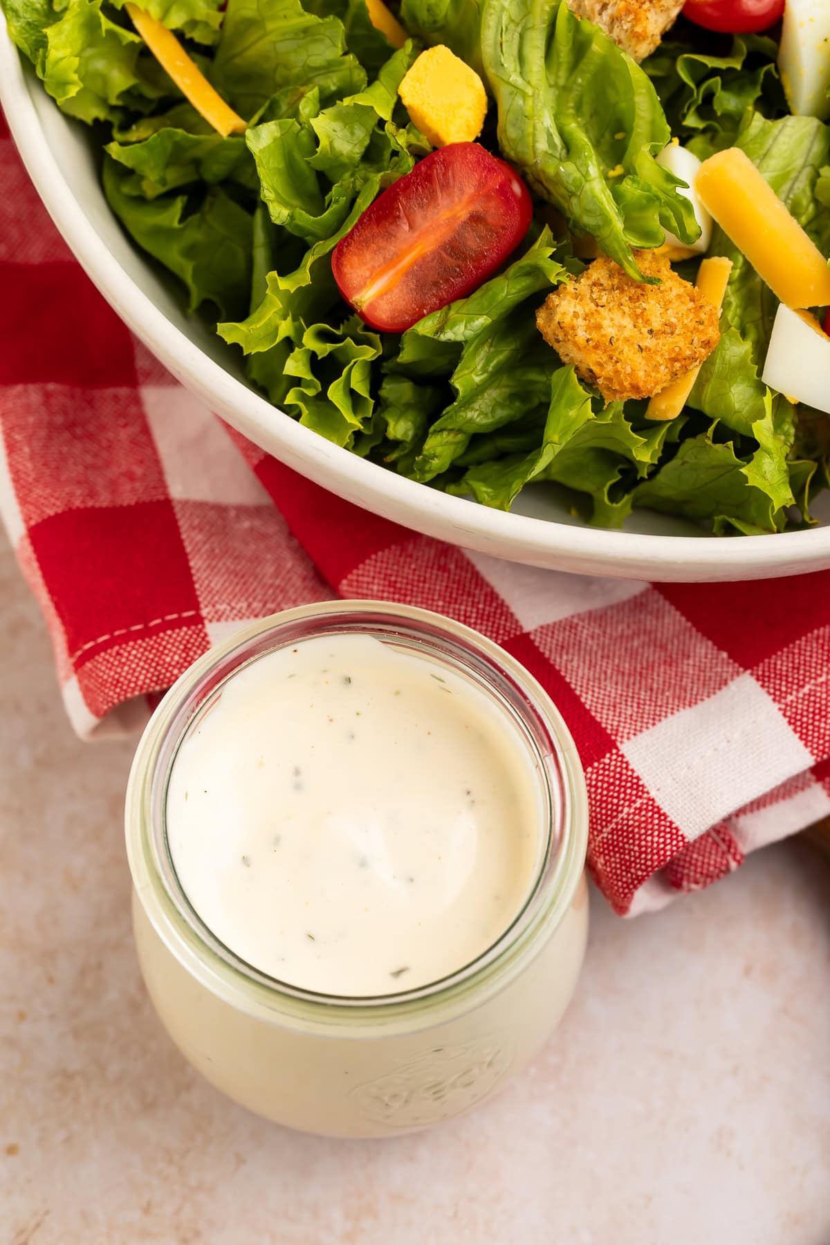Top down view of a creamy ranch dressing in a glass jar next to a salad sitting on a gingham napkin. 