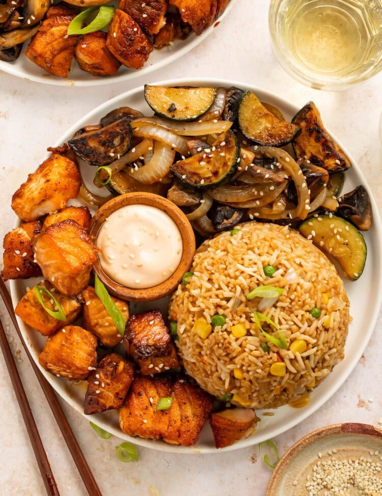 Bowl of hibachi salmon, mixed vegetables including mushroom, zucchini and onions, served with a mound of fried rice and a small wooden pot of creamy dipping sauce.