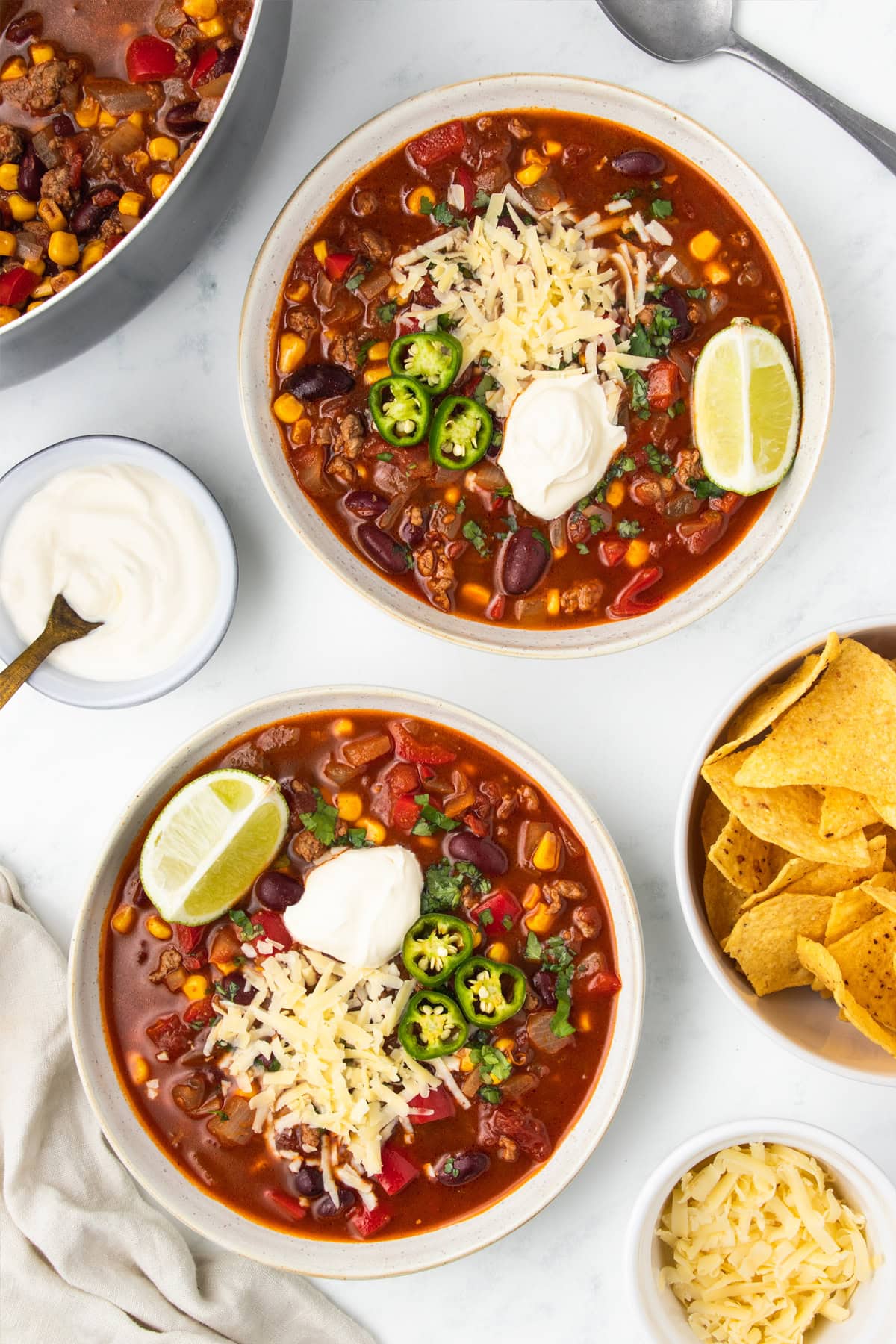 Two white bowls of chili surrounded bowls with fixings of sour cream and tortilla chips.