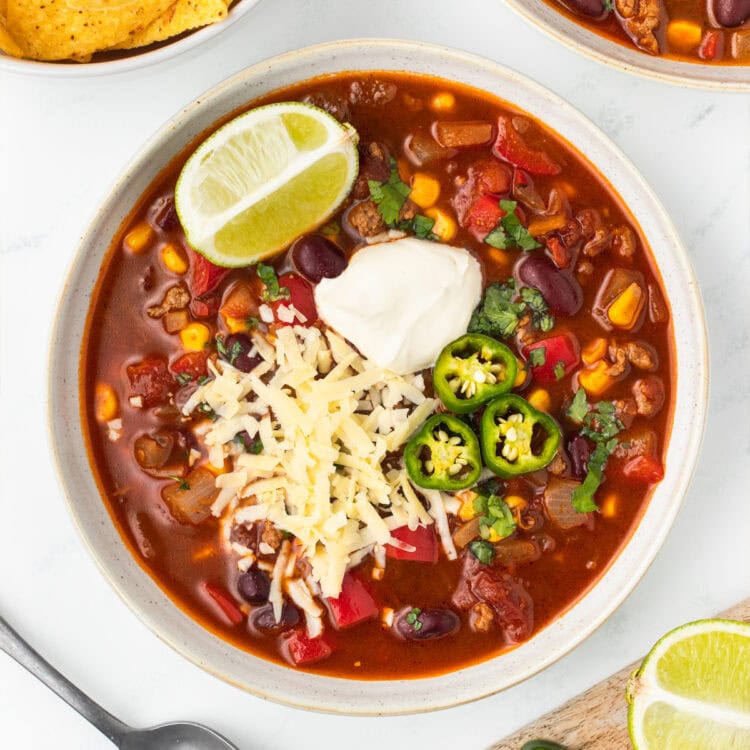 Bowl of chili topped with sour cream, shredded cheese, sliced jalapeños and a lime wedge.