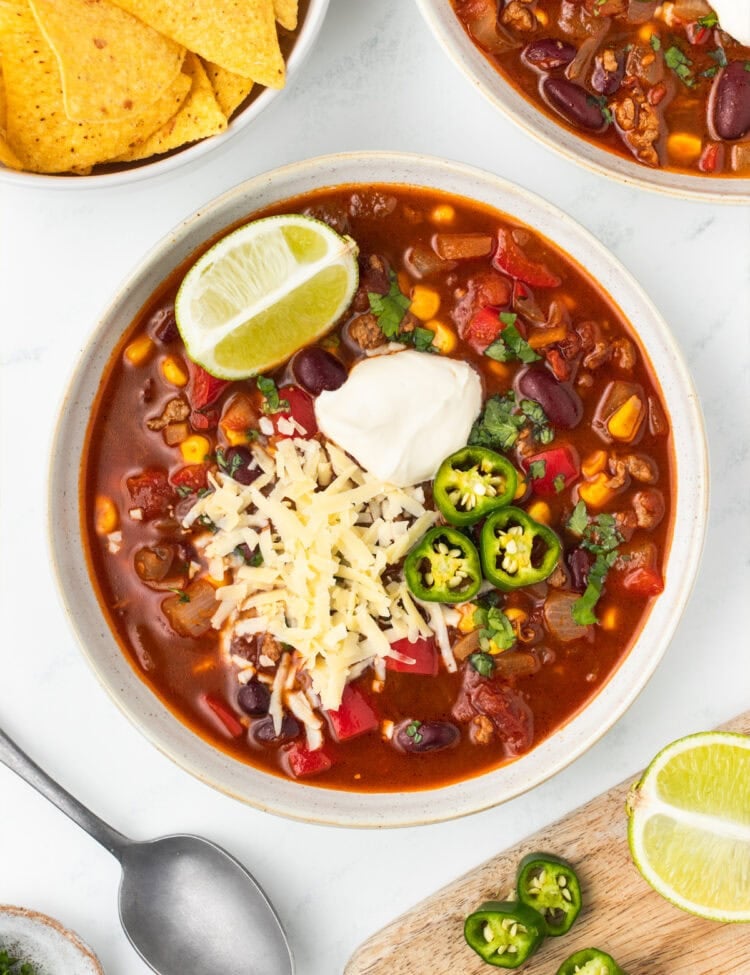 Bowl of chili topped with sour cream, shredded cheese, sliced jalapeños and a lime wedge.