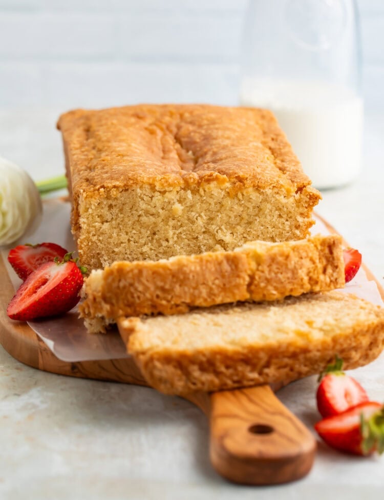 Sliced vegan pound cake on a wooden board with fresh flowers and sliced strawberries just visible off to the side.