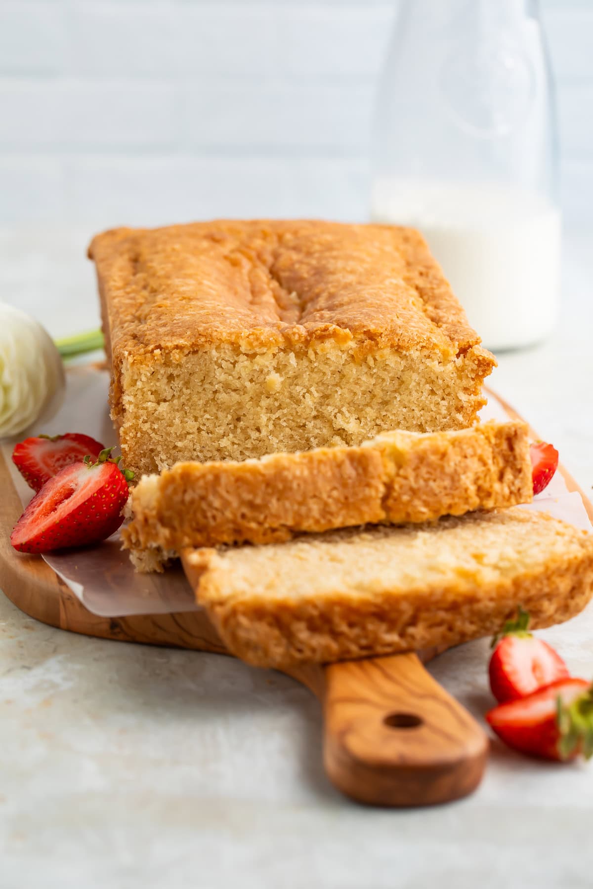 Sliced vegan pound cake on a wooden board with fresh flowers and sliced strawberries just visible off to the side.