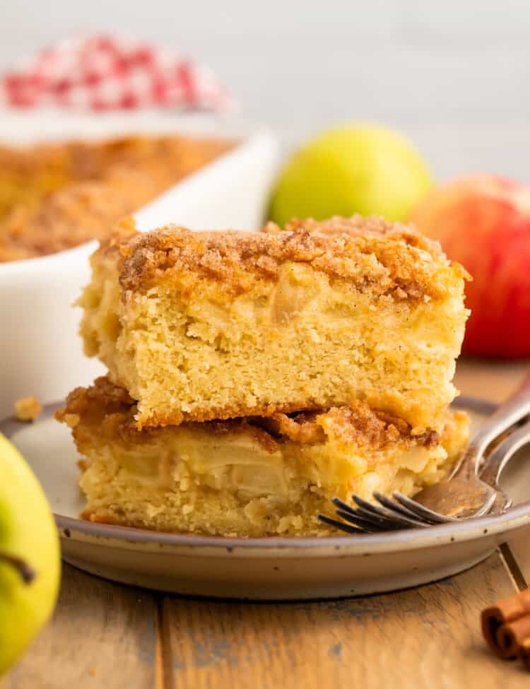 Slice of apple cake with a cinnamon sugar topping on a grey place, with the pan of cake and fresh apples in the background.