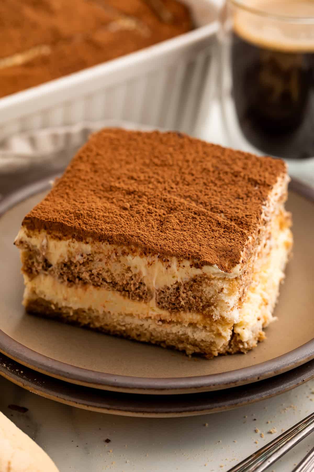 Slice of tiramisu on a grey plate with a cup of coffee and the pan of dessert visible in the background.
