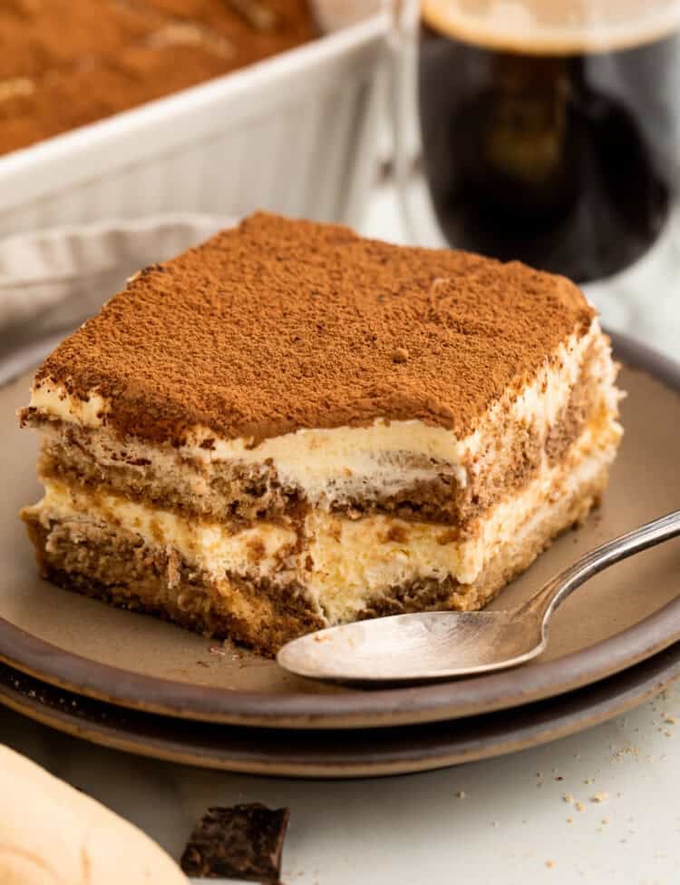 Slice of tiramisu on a plate with a spoon where a spoonful has just been taken out of the corner.