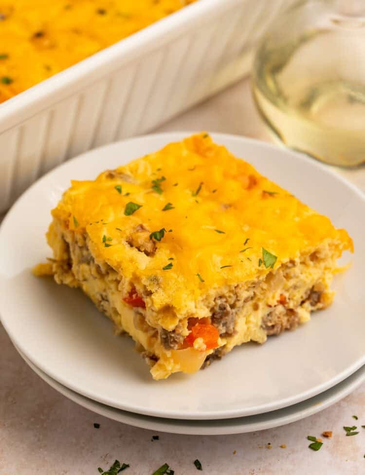 Slice of egg bake on a white plate with the casserole in the background in a white dish.