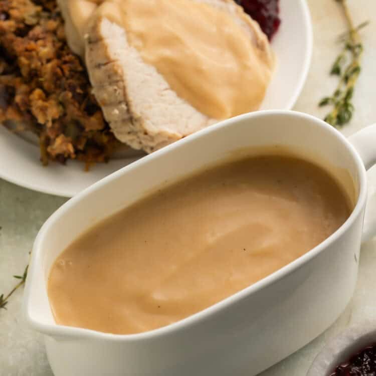 White gravy boat of gravy with gravy-covered turkey and stuffing on a white plate in the background.