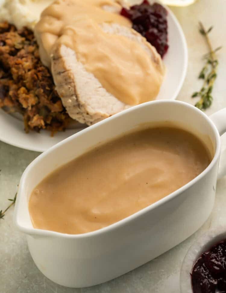 White gravy boat of gravy with gravy-covered turkey and stuffing on a white plate in the background.