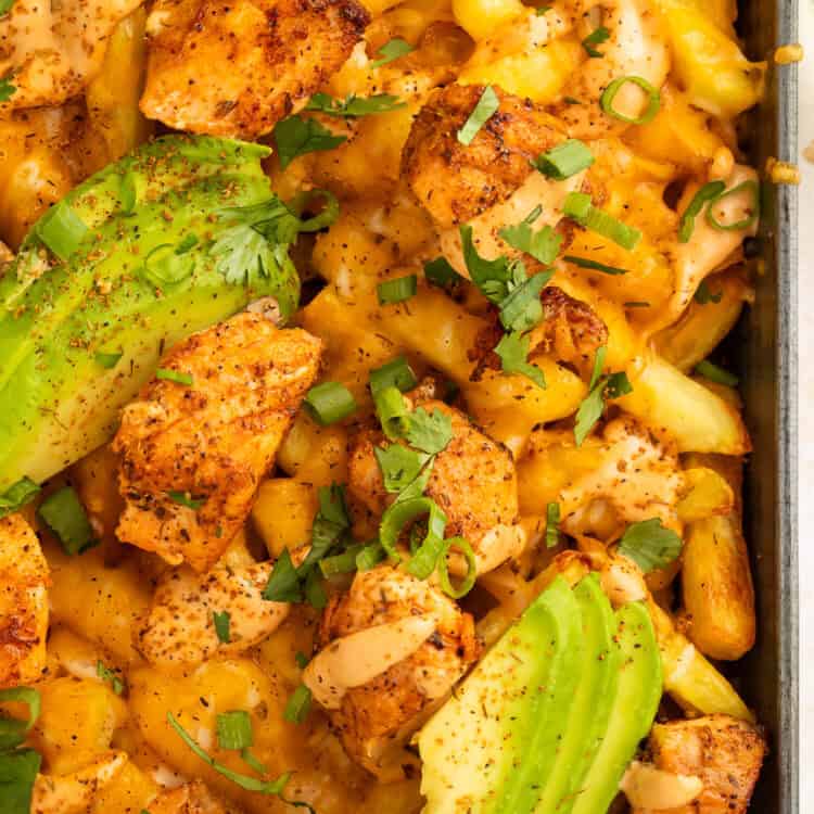 Close up of a tray of French fries topped with pieces of blackened salmon, sliced avocado and a creamy sauce.