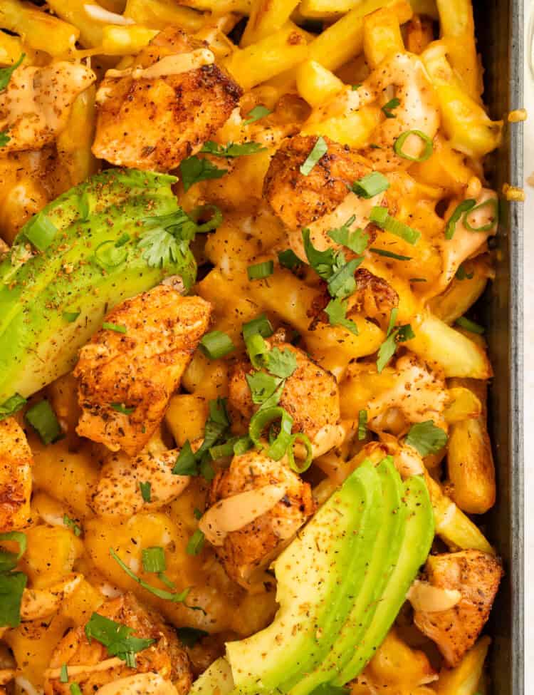 Close up of a tray of French fries topped with pieces of blackened salmon, sliced avocado and a creamy sauce.