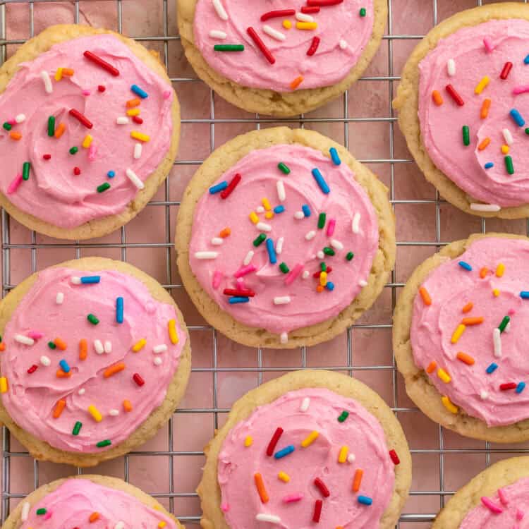Pink frosted sugar cookies with rainbow sprinkles on a cooling rack.