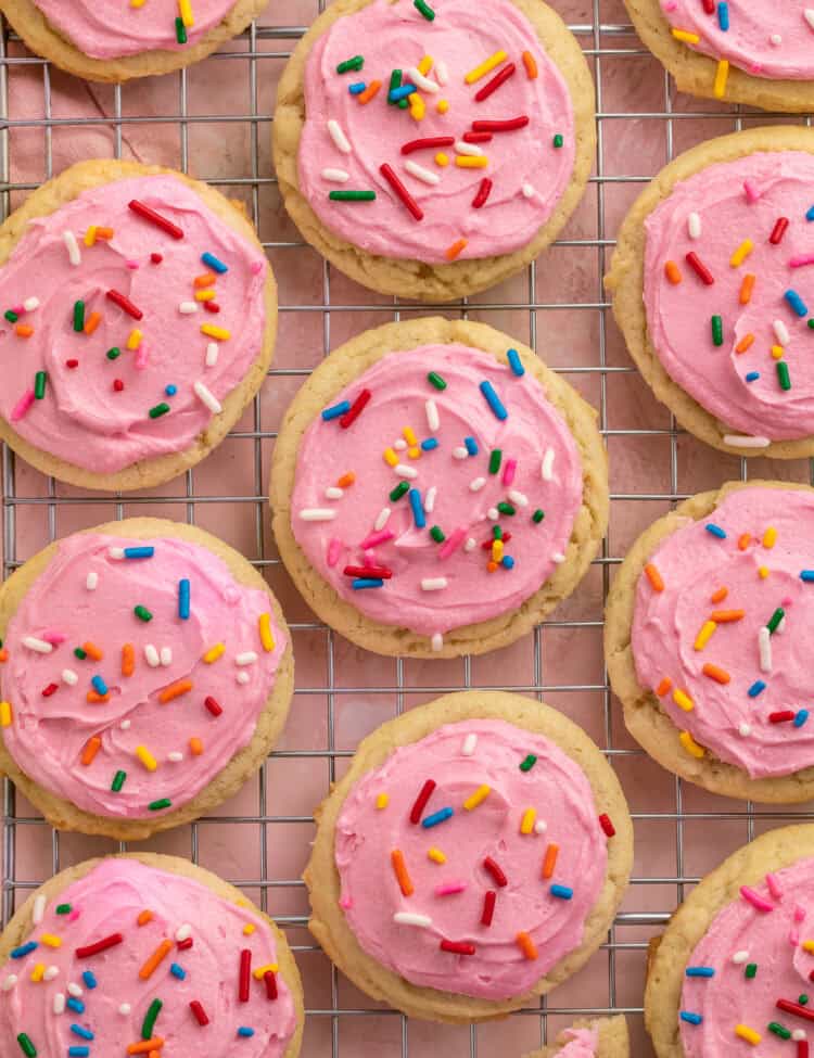Pink frosted sugar cookies with rainbow sprinkles on a cooling rack.