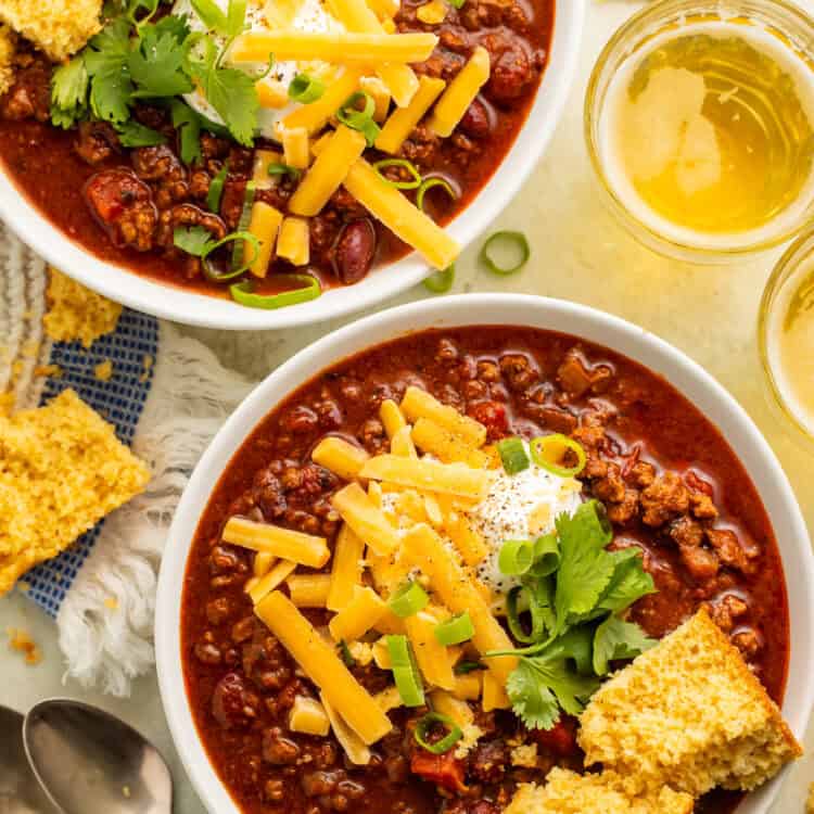 Two white bowls of chili topped with cornbread, green onions, grated cheese and sour cream.