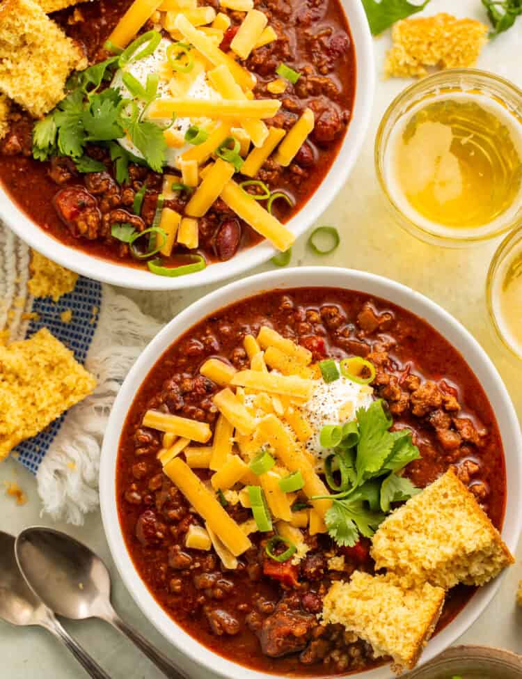 Two white bowls of chili topped with cornbread, green onions, grated cheese and sour cream.