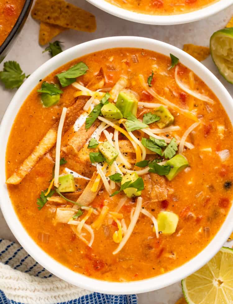 Close up of a white bowl of tortilla soup topped with avocado, shredded cheese and cilantro.