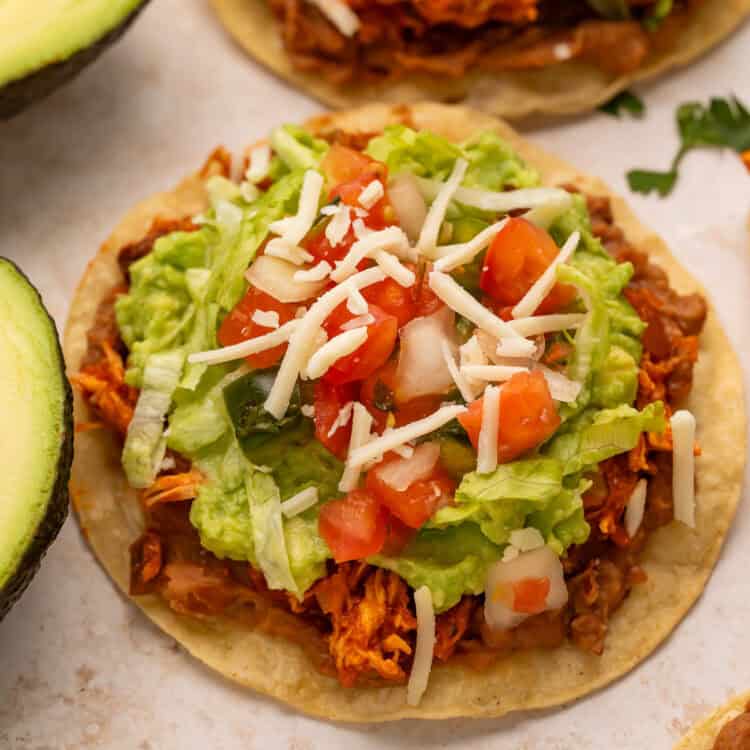 Close up of a chicken tostada topped with shredded lettuce, guacamole, salsa and shredded cheese.