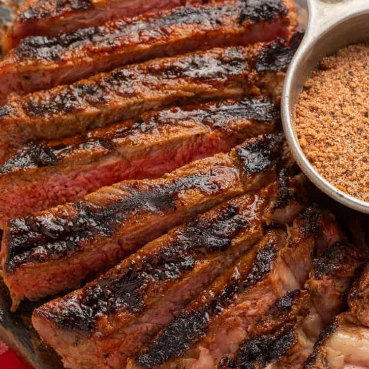 Close up of a sliced steak next to a metal measuring cup of steak seasoning.